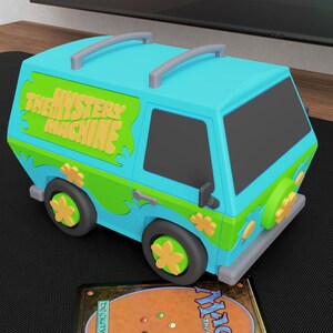 May include: A light blue and green toy van featuring the text THE MYSTERY MACHINE on the side, with flower-shaped wheel covers and roof rails. Positioned in front of the van is a rectangular Magic The Gathering trading card with a brown back.