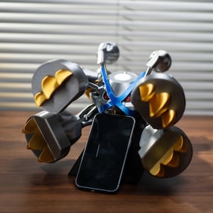 May include: A futuristic-looking phone stand in silver, gray, and yellow, holding a black smartphone. The stand has a mechanical design with circular elements and a blue accent. The phone is positioned upright.