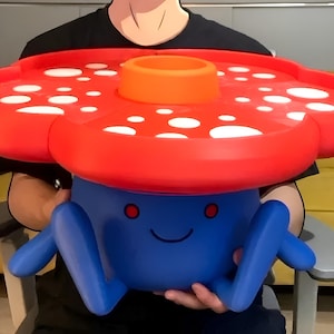 May include: A blue plastic creature-shaped stool with a large, flat, red mushroom-cap top featuring white circular spots and a central orange ring. The blue base has two protruding legs, two small red circular eyes, and a simple curved black line for a mouth.