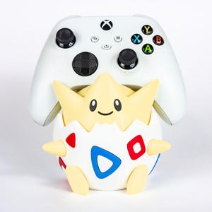 Op de afbeelding: Een witte Xbox-controller staat achter een Togepi-figuur. De Togepi-figuur is geel, wit, rood en blauw. De controller heeft zwarte joysticks en gekleurde knoppen.