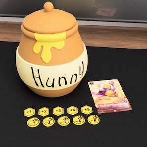 May include: A tan honey pot with a lid, featuring a yellow honey drip design and the word "Hunny" written in black. Beside it lies a rectangular trading card depicting a cartoon bear and ten yellow hexagonal tokens marked with numbers.
