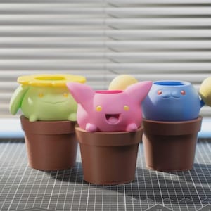 Puede incluir: Tres macetas de estilo caricatura. Una es verde claro con una parte superior de flor amarilla, otra es rosa con orejas y la última es azul con una bola amarilla. Todas están en macetas marrones.