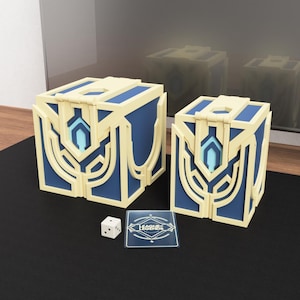 Könnte beinhalten: Zwei blaue und goldfarbene dekorative Boxen mit einer Karte und einem Würfel. Die Boxen haben ein geometrisches Design mit einem hellblauen Akzent. Die Karte hat den Text "League of Legends". Der Würfel ist weiß mit schwarzen Punkten.