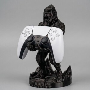 May include: A black gorilla-shaped controller stand holding a white and black PlayStation 5 controller. The stand is detailed with textured fur and a rocky base, set against a gray background. A unique gaming accessory.