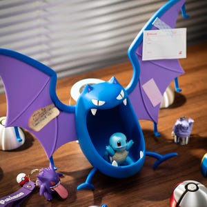 May include: A blue cartoon figure with purple wings, holding a small Squirtle figurine. Other Pokemon-themed figurines and keychains are scattered around, including a purple Gengar and various Poke Balls. The scene is set on a wooden surface.