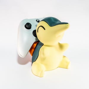 Puede incluir: Una figura de Cyndaquil amarilla y verde azulado sosteniendo un mando de Xbox blanco. La figura tiene una cara sonriente y está en posición sentada. El mando tiene botones negros y un joystick negro.