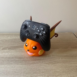 Op de afbeelding: Een zwarte gamecontroller rust op een oranje en gele standaard in de vorm van een personage. De controller heeft een sterontwerp en meerdere knoppen. De standaard heeft een lachend gezicht en puntige oren. De items staan op een houten oppervlak.