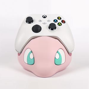 Peut inclure: Une manette Xbox blanche repose sur un support de manette rose de style dessin animé. Le support présente un visage rose avec de grands yeux de couleur sarcelle. La manette a des boutons noirs et des boutons d'action colorés.
