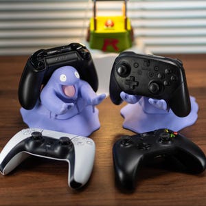 Puede incluir: Cuatro mandos de videojuegos, incluido un mando de PlayStation 5, descansan sobre una superficie de madera. Los mandos están emparejados con soportes morados con forma de personajes de dibujos animados. Hay una pequeña estructura amarilla y verde en el fondo.