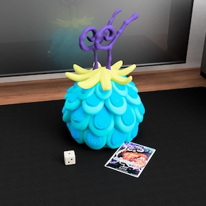 May include: A spherical blue fruit-shaped object with a textured scale pattern, topped with yellow leaves and two purple swirling stems. Beside it sits a small white six-sided die and a rectangular trading card featuring a character illustration.