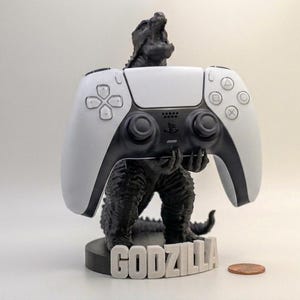 May include: A black and white Godzilla figure holding a white and black PlayStation 5 controller. The base of the figure reads "GODZILLA" in white letters. A penny is next to the base.