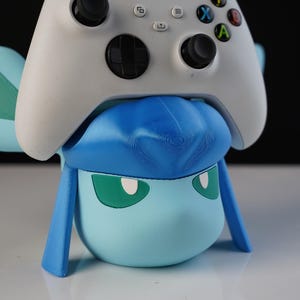 May include: A light blue and white controller holder shaped like a cartoon character, with a white Xbox controller resting on top. The character has green eyes and blue accents. The background is black.