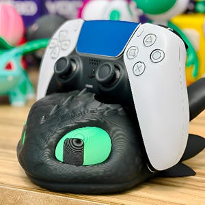 Könnte beinhalten: Ein schwarzer und weißer PlayStation 5-Controller liegt auf einem schwarzen Controller-Ständer im Drachen-Design. Der Controller hat ein blaues Mittelteil und weiße Seiten mit schwarzen Joysticks. Der Drachenständer hat grüne Augen.