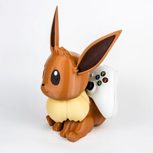 Puede incluir: Una figura marrón de Eevee con orejas grandes y un volante de color crema, sosteniendo un mando de Xbox blanco. El mando tiene botones de colores. La figura es un artículo decorativo para jugadores.