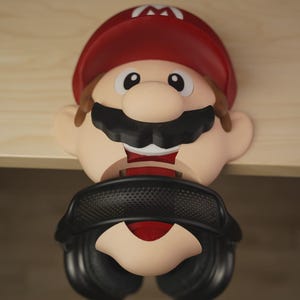 May include: A novelty headphone stand in the shape of the character Mario. The stand has a red hat with a white "M", a black moustache, and a smiling face. Headphones rest on the character's face, with the earcups on either side.