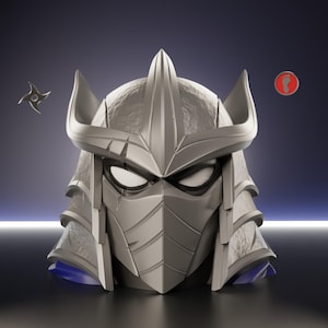 Op de afbeelding: Een gedetailleerde, zilverkleurige sculptuur van Shredders helm uit Teenage Mutant Ninja Turtles. De helm heeft een prominente centrale punt, hoekige details en een gezichtsmasker. Een ninja-ster en een rood-wit symbool zijn ook aanwezig.