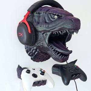 May include: A purple and black dinosaur head wall mount with a pair of black and red headphones. Below are two game controller holders, one white and one black, designed to look like dinosaur claws.