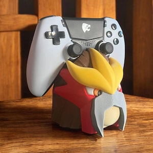 May include: A white and black gaming controller resting on a 3D-printed figure with red, yellow, brown, and gray accents. The figure has a stylized design, and the controller features a directional pad and buttons.