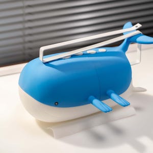 May include: A blue and white whale-shaped speaker with a white handle. The speaker has a rounded body, fins, and a tail. It has three white buttons on top and a small black dot on the side.
