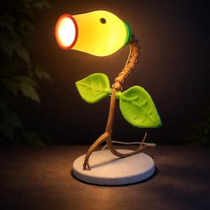 Puede incluir: Una lámpara de escritorio caprichosa con forma de planta de dibujos animados. La lámpara presenta una cabeza amarilla brillante con un borde rojo, detalles verdes y dos hojas verdes. El tallo es marrón y la base es un círculo gris claro.
