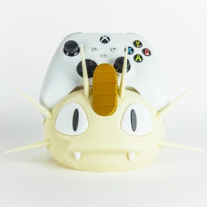 May include: A cream-colored, 3D-printed Meowth-themed controller holder with a gold accent on top. A white Xbox controller rests on top of the holder. The controller has black joysticks and colored buttons.