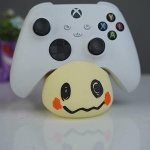 Op de afbeelding: Een witte Xbox-controller rust bovenop een gele Mimikyu-figuur. De controller heeft zwarte joysticks en knoppen met gekleurde X-, Y-, A- en B-knoppen. De Mimikyu-figuur heeft een zwart getekend gezicht met oranje wangaccenten.