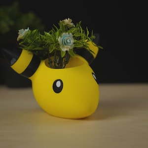 Puede incluir: Una maceta amarilla de estilo caricatura con rayas y orejas negras, que contiene pequeñas plantas artificiales con flores azules y blancas. La maceta tiene ojos negros.