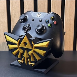 Puede incluir: Mando Xbox negro sobre un soporte decorativo negro y dorado. El soporte presenta un emblema dorado con un diseño de triángulo. El mando tiene botones de colores y controles direccionales.