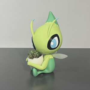May include: A green and yellow figurine of a cartoon character holding a small white eggshell planter with greenery. The figurine has large blue eyes, small wings, and blue antennae. The figurine is sitting on a dark gray surface.
