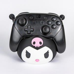 May include: A black Nintendo Switch Pro controller docked in a black and white character stand. The stand features a white face with black eyes, a pink nose, and a pink skull icon on the forehead. The controller has standard buttons and joysticks.