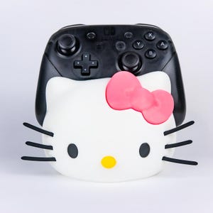 Op de afbeelding: Een zwarte Nintendo Switch Pro-controller met een witte Hello Kitty-gezichtshoes. De hoes is voorzien van een felroze strik, een gele neus, zwarte ogen en drie zwarte snorharen aan elke kant van het witte gezicht, die de controller deels bedekken.