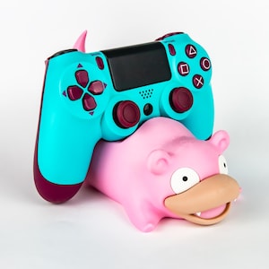Op de afbeelding: Een turquoise en bordeauxrode PlayStation 4-controller rust op een roze Slowpoke-figuur. De controller heeft de standaard knoppenindeling met een zwart touchpad. De Slowpoke-figuur heeft een glimlachende uitdrukking.