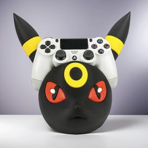 Puede incluir: Un soporte para mando de PlayStation 4 con temática Umbreon negro y amarillo con un mando blanco. El soporte tiene ojos rojos y un anillo amarillo. El mando es blanco con botones negros.