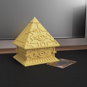 May include: A golden-yellow pyramid-shaped box with intricate designs and the Eye of Wdjat symbol. The box is on a black surface, next to a trading card. The pyramid box is a collectible item.
