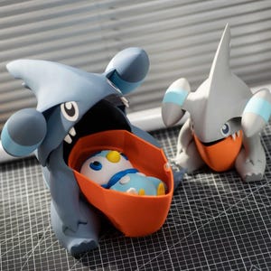 Puede incluir: Dos figuras de Pokémon. Una es una criatura grande, azul-gris con la boca abierta que contiene un interior naranja y una figura azul y blanca más pequeña. La otra es una figura gris con detalles en naranja.