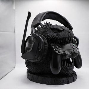 Puede incluir: Soporte negro para auriculares y mando de videojuegos con temática de Godzilla. El soporte presenta una escultura detallada de la cabeza de Godzilla, sosteniendo un mando negro en su boca. Un auricular negro para juegos descansa sobre la cabeza.