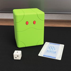 Könnte beinhalten: Eine limettengrüne Kartenaufbewahrungsbox mit zwei roten kreisförmigen Akzenten, einem weißen Würfel und einer Karte mit dem blauen Text "GUNDAM CARD GAME". Die Box ist würfelförmig und steht auf einer schwarzen Oberfläche.