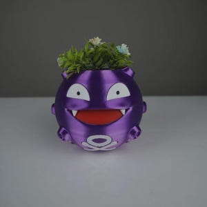 Puede incluir: Una maceta redonda morada con forma de personaje de dibujos animados con ojos, dientes y boca blancos. La maceta está llena de plantas artificiales verdes y blancas. La maceta tiene un diseño blanco en la parte inferior.