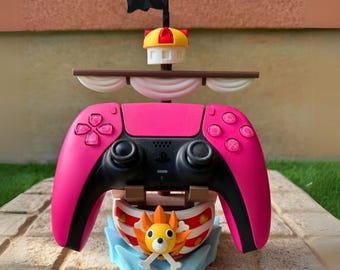 May include: A pink and black video game controller with a pirate ship-themed stand. The stand features a mast with a black flag, a yellow and red top, and a cartoon dog figure. The controller has pink buttons and a black center.