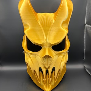 Peut inclure: Un masque intégral doré avec un design en forme de crâne. Le masque présente de grandes cornes incurvées, des orbites et une bouche dentelée. Le masque est fait d'un matériau texturé, probablement pour un costume ou à des fins décoratives.