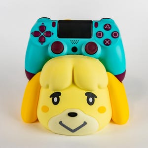 May include: A teal and maroon video game controller rests atop a yellow, cartoon-style head with black eyes and a black nose. The controller has a black center panel and colored buttons. The head has yellow ears and a smiling expression.