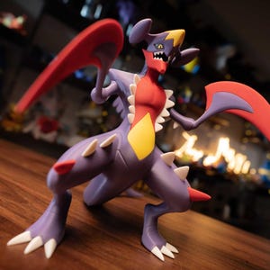 Puede incluir: Figura detallada de Garchomp, un personaje de Pokémon. La figura es principalmente morada con acentos rojos, amarillos y blancos. Tiene grandes apéndices rojos en forma de cuchilla y está sobre una superficie de madera.