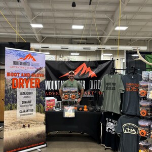 Op de afbeelding: Een stand op een beurs met een man die voor een banner staat met de tekst "Mountain Hunter Outdoors Adventure Starts Here Boot and Glove Dryer". De banner adverteert een droger die ultralicht, duurzaam, waterbestendig is en een jaar garantie heeft. De stand heeft ook een tafel met verschillende producten, waaronder T-shirts, hoeden en andere outdooruitrusting.
