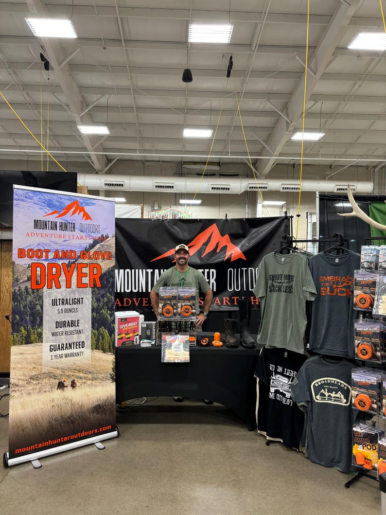 Op de afbeelding: Een stand op een beurs met een man die voor een banner staat met de tekst "Mountain Hunter Outdoors Adventure Starts Here Boot and Glove Dryer". De banner adverteert een droger die ultralicht, duurzaam, waterbestendig is en een jaar garantie heeft. De stand heeft ook een tafel met verschillende producten, waaronder T-shirts, hoeden en andere outdooruitrusting.