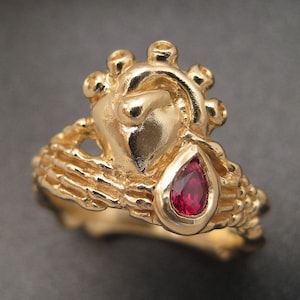 Puede incluir: Un anillo de oro con un diseño en forma de corazón y una piedra preciosa roja. El anillo está intrincadamente detallado y tiene una superficie texturizada.