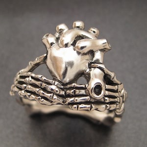 May include: A silver ring featuring a detailed anatomical heart design held by two skeletal hands. The ring is made of metal and has a textured finish.