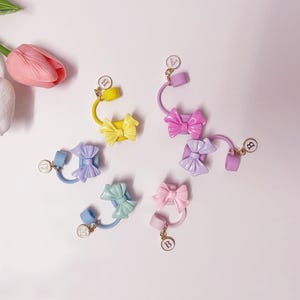 Pode incluir: Cinco elásticos de cabelo coloridos com laços e pingentes dourados. Os laços são em tons de rosa, roxo, azul e amarelo. Os pingentes são redondos e têm a letra "B" neles.