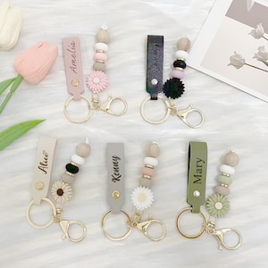May include: Six personalized keychains with gold-colored hardware. Each keychain features a leather strap with a name, decorative beads, and a flower charm. The names include "Amelia", "Alice", "Kenny", and "Mary". The keychains have various color schemes.