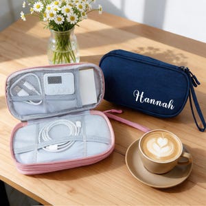May include: A pink zippered organizer case with internal mesh pockets holding white charging cables and a small digital device, alongside a closed navy blue zippered case with the name Hannah printed in white script on the front.