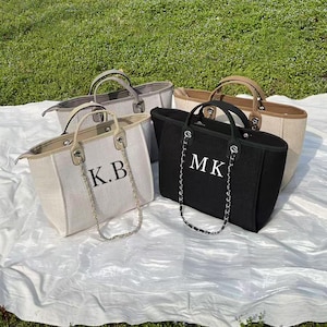 May include: Three canvas tote bags with chain straps. The bags are in beige, black, and brown. The beige bag has the initials "K.B." embroidered on the front. The black bag has the initials "MK" embroidered on the front. The brown bag has no visible embroidery.